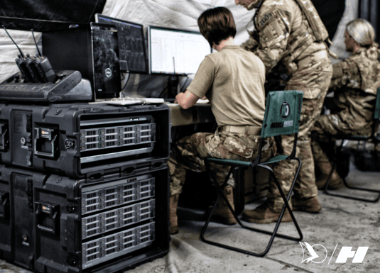 Military personnel operating a tactical command center inside a field tent, working on multiple computer screens. Stacked rugged rack-mounted server cases provide secure data processing and communication support.