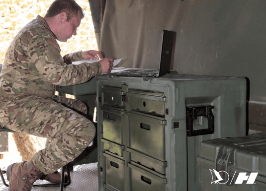 Military personnel working on a laptop inside a field tent, using a rugged deployable workstation with multiple storage drawers for secure equipment organization.
