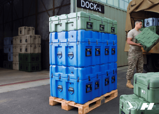 Military personnel loading a rugged green storage case onto a transport vehicle. Stacks of blue and green heavy-duty cases are organized on a Euro pallet for secure equipment transport and deployment.