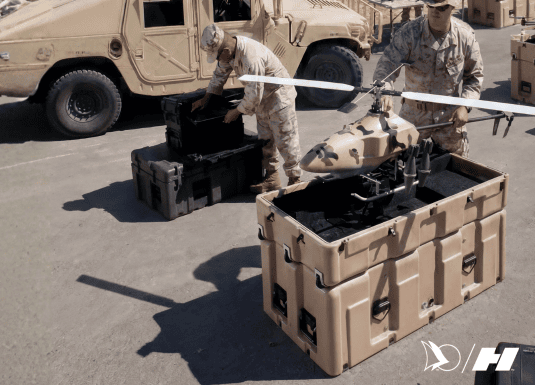 Military personnel assembling a tactical unmanned aerial vehicle (UAV) on top of a rugged beige transport case. Military vehicles and additional equipment are visible in the background.