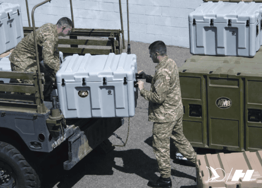 Military personnel in camouflage uniforms loading a rugged white storage container onto a transport vehicle. Additional heavy-duty cases and equipment are stacked nearby for deployment.