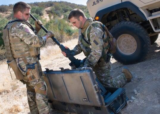Two tactical military personnel in desert camouflage handling a rifle near an open rugged hard case in an arid environment. A military vehicle is parked nearby, indicating a field operation or deployment.